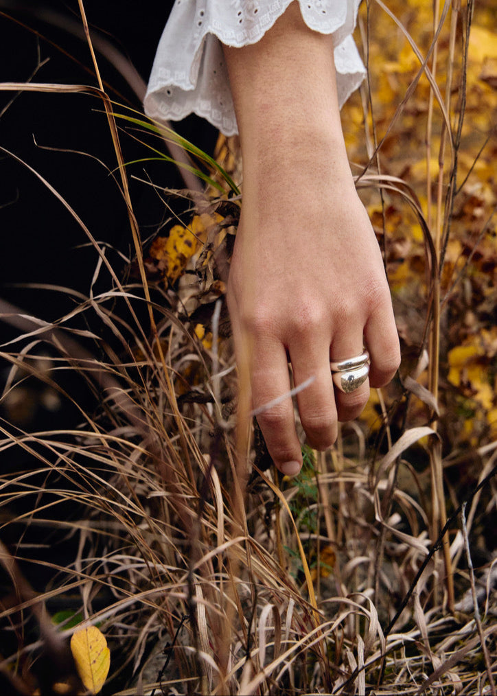 Detailed view of the Jooni Ring stacking it on the models finger with the ingrid ring and touching the grass surrounding her. The photo is focusing on its 5mm width and the reflective quality of the 925 sterling silver.