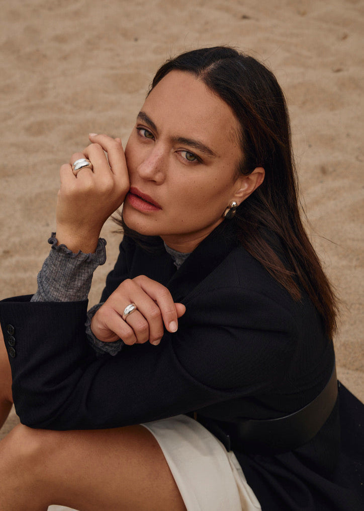 Close-up of a model wearing the Nilou Ring while sitting on the beach, emphasising its bold design and comfortable fit. she has it stacked with the smaller sister Jooni ring. The picture is very serious but also showing the versatile look of the ring